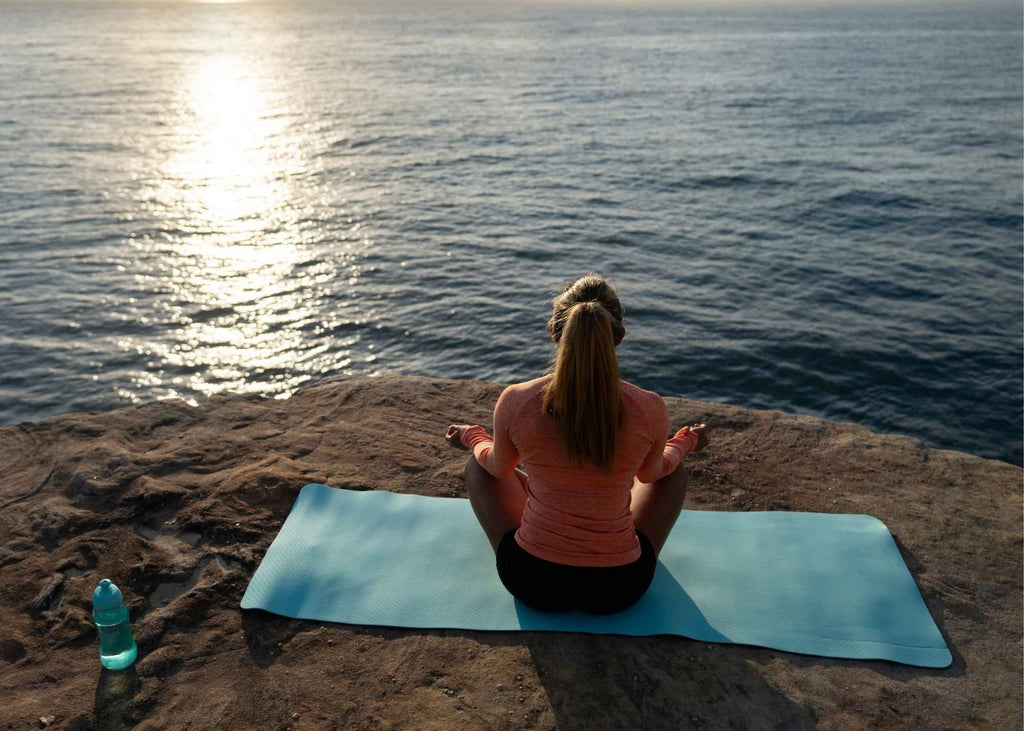 Woman meditating as she sits on yoga mat by sea, drink bottle by her side.. 